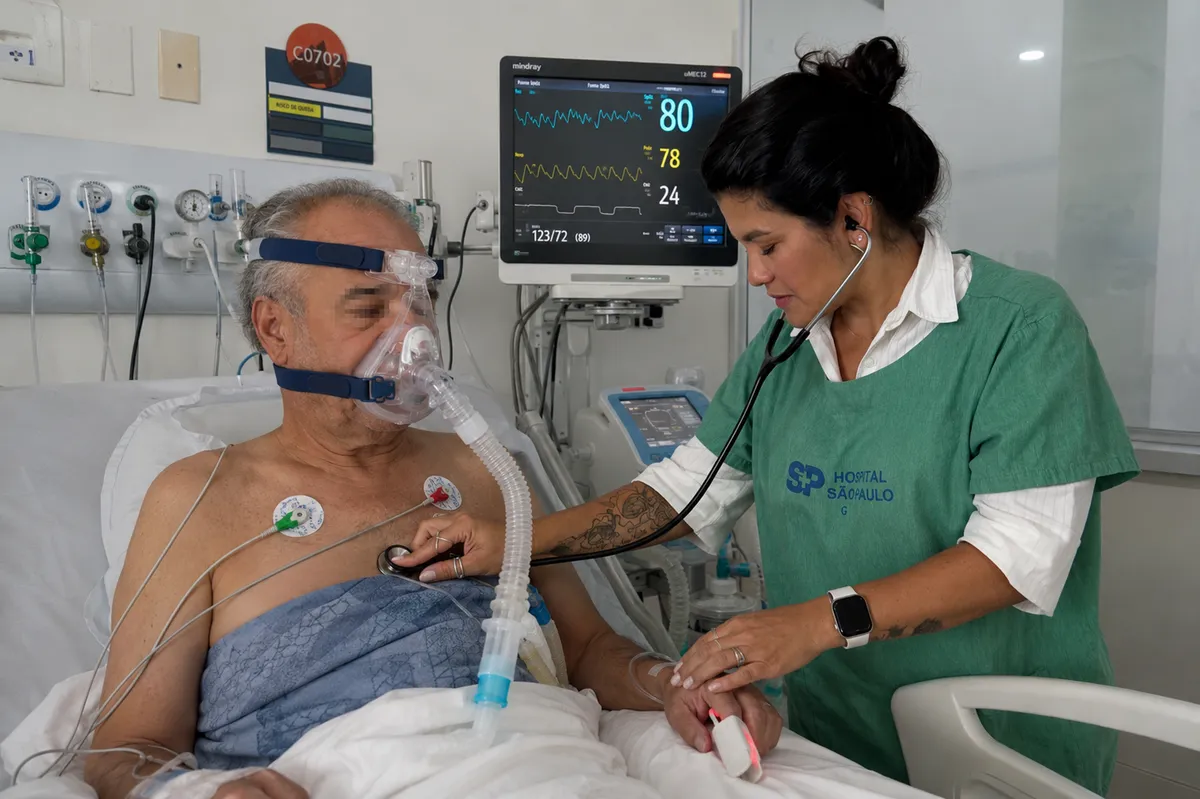 Paciente na UTI em ventilação não invasiva, com a máscara ajustada, enquanto a fisioterapeuta ausculta o tórax e acompanha o monitor.